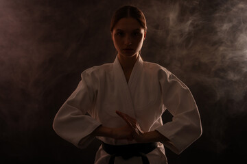Woman in uniform practicing karate on black background with smoke