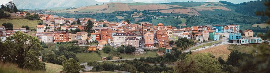 Obraz premium Telephoto panoramic view of Tineo town in Asturias, colorful buildings and red tile roofs on green hills and farmland, Camino de Santiago Primitivo route landscape, cloudy mountain backdrop