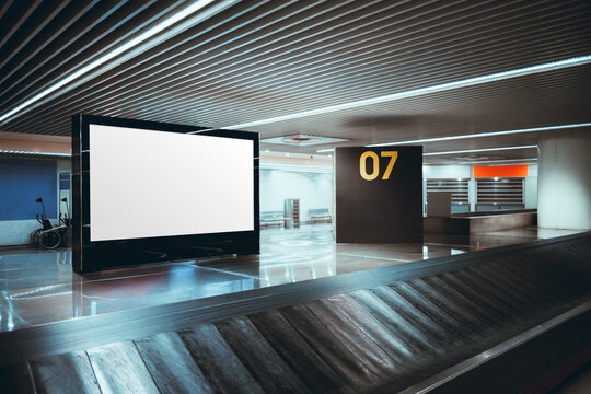 Empty horizontal billboard mockup beside airport baggage claim conveyor, modern terminal interior with ceiling strip lights and carousel number 07, blank advertising display with copy space