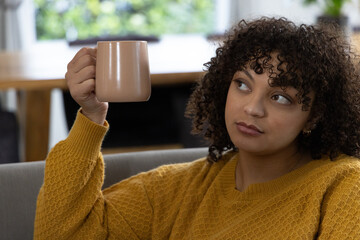 African American woman sitting on sofa holding beige mug near face wearing mustard-yellow sweater