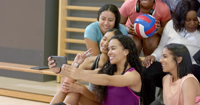 Female volleyball teammates positioning smartphone for selfie while holding volleyball on bleachers