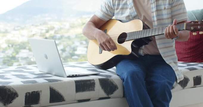 Seated man is placing left hand on neck, causing strumming acoustic guitar for practice near laptop