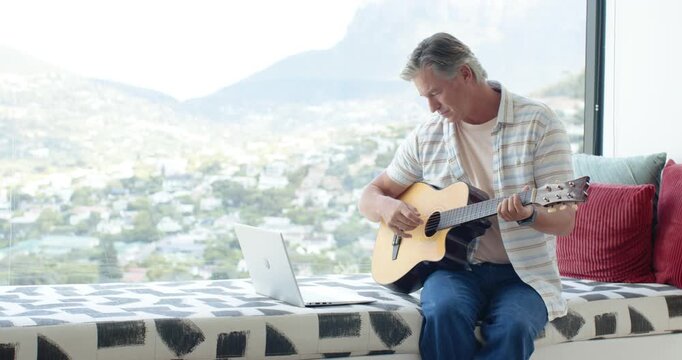 Mature male sitting on bench by window following laptop tab while playing guitar, copy space