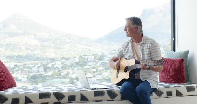 Man sitting by window, glancing out, strumming guitar and checking laptop, copy space