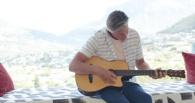 Man is settling acoustic guitar on lap, tuning and playing for relaxation on bench by valley