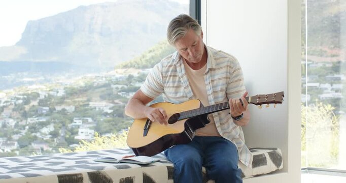 Mature man sitting by tall window, glancing at notebook, tuning and practicing chords on guitar