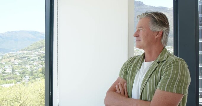 Mature man standing by window reacting to sunlit valley, wearing green shirt, smiling, copy space