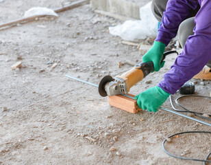 A construction worker using an electric angle grinder to cut a threaded metal rod on a brick support.