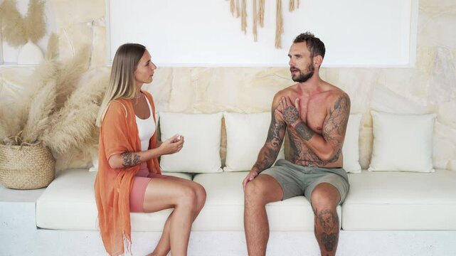 Female physiotherapist talking to her male client in a luxury house setting with neutral colour tones. They are both sitting on a couch 