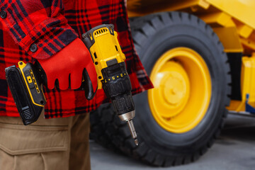 Close up of construction worker in red checkered jacket and protective gloves holding yellow cordless drill with battery pack near large wheel of heavy machinery. Concepts of construction services.