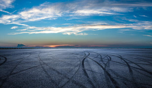 Wide open asphalt race track featuring black tire burnout marks under a dramatic sunset sky