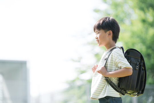 学校の校舎と緑を背景に校庭でランドセルを背負って走る少年の(男の子)男子の横顔、逆光の未来や希望を見つめるスタートするイメージ　コピスペースあり