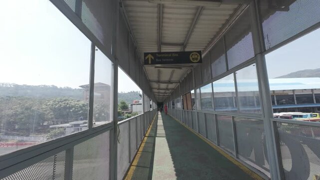 A long, enclosed pedestrian bridge or skywalk with glass windows and metal mesh, featuring a directional sign pointing towards the bus terminal. 