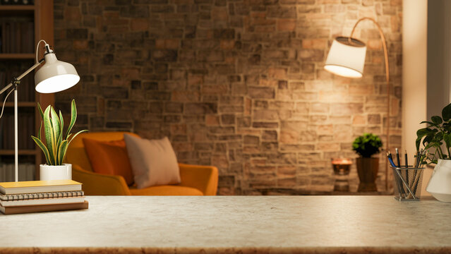 Books and potted plant under lamp on marble counter table with sofa chair in dim lit brick wall room