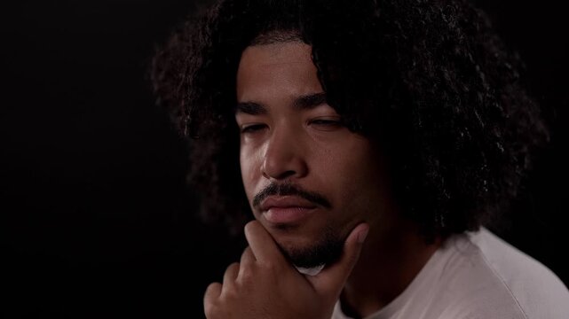 Portrait of a thoughtful young African-American man with curly hair and goatee posing with his hand on his chin against a black background