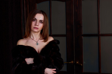 Obraz premium A woman in a black fur coat, draped over her naked body, stands at the glass door. She has black stockings on her feet.