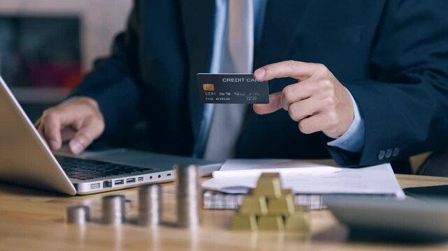 Businessman holding credit card and using laptop for online gold trading. Investor checking credit score and analyzing financial risk for investment on office desk. 4K footage.