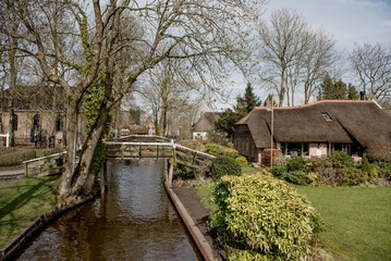 Obraz premium Giethoorn, Netherlands, April 08 2018: Scenic view of a charming canal with a wooden bridge and traditional thatched-roof houses surrounded by lush greenery
