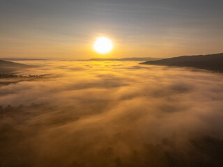 Fototapeta premium A cinematic drone aerial view of sunrise casting golden light over a misty mountain valley, with a sea of fog drifting through the landscape and distant peaks on the horizon.