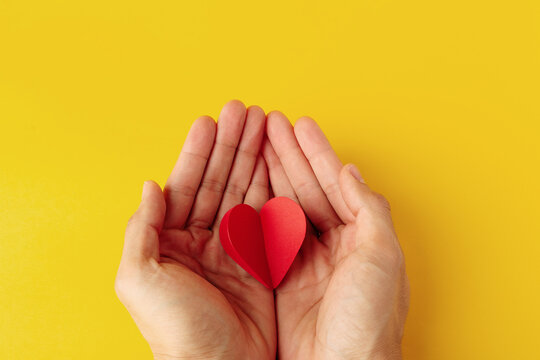 Two hands gently cupping a red paper heart on a yellow background. Represents empathy, protection, mental health support, and shared love. Top view with ample copy space for text and design.