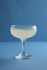 Elegant frosted coupe glass filled with pale yellow liquid stands on a blue surface against a muted blue background