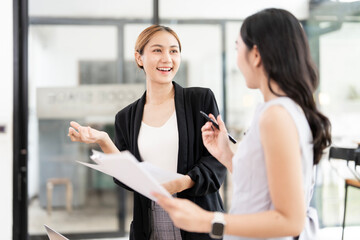 Obraz premium Two professional Asian businesswomen discussing project reports and collaborating in a bright, modern office environment, showcasing teamwork and effective communication.