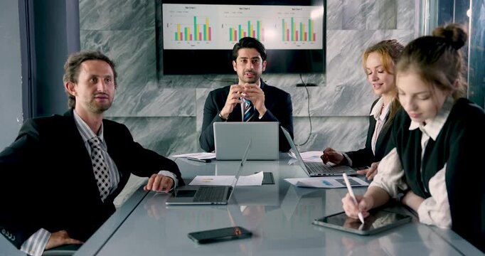 Multicultural team discussing new business ideas, businessman speaking while team reviews digital documents and charts on devices, collaborating in modern office workspace environment