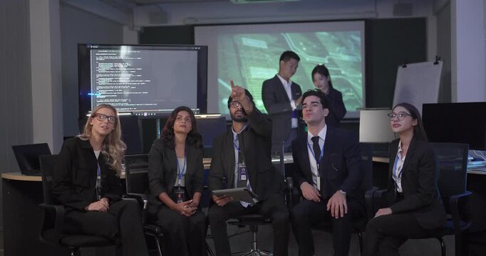 Diverse business team gathers for roundtable presentation in tech office, with a presenter leading discussion as colleagues review documents and screens. Collaboration, training, strategic planning
