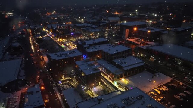 Aerial night view of American city during snowfall, glowing street lights and illuminated buildings. Calm winter atmosphere as snow falls over rooftops and streets. Wide shot.