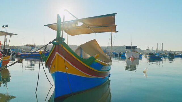 Colorful luzzu fishing boats floating on calm water at sunrise in Marsaxlokk harbor, Malta.