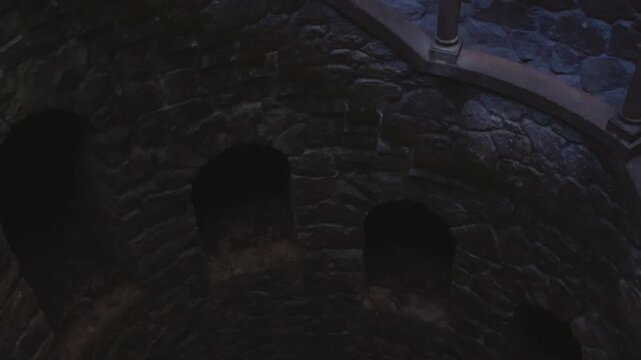 Cinematic tilt down into the historic Initiation Well, revealing ancient stone walls, carved openings, and a deep spiral descent into shadowed underground space.