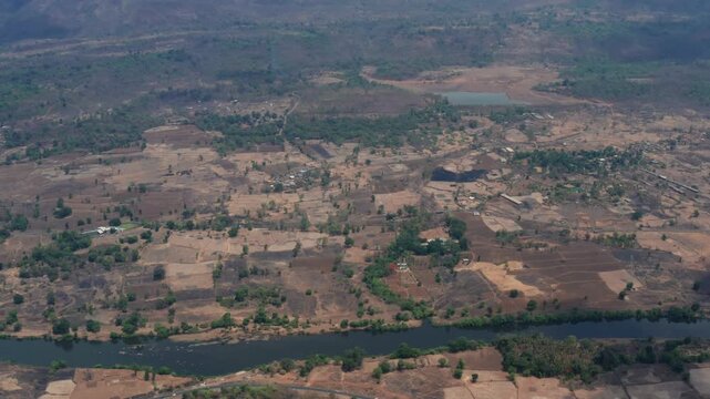 Slow drone ascends from winding Amba River revealing expansive arid farmlands, sparse vegetation, small ponds, roads, and distant hills in Pali Raigad region Maharashtra.