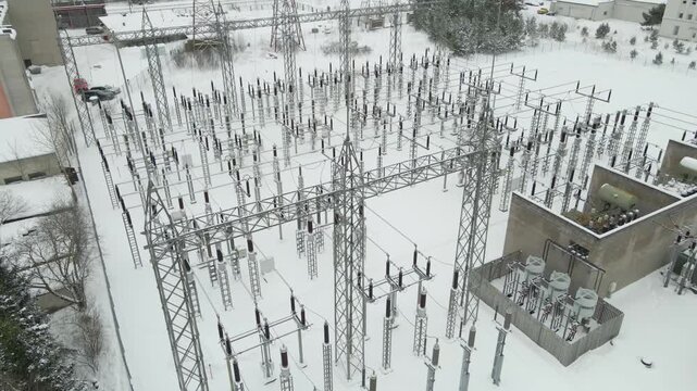 Wide aerial orbit over a large electrical substation in extreme winter conditions, showing snow covered insulators, transformers, and power lines under clear cold daylight.