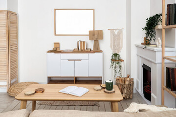 Naklejka premium Interior of living room with commode, coffee table, houseplants, fireplace and firewood in wicker basket near white wall