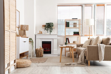 Naklejka premium Interior of living room with sofa, shelving unit, coffee table, fireplace, commode and firewood in wicker basket near window