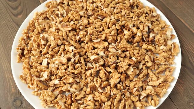 Shelled and chopped walnut kernels rotating on a white plate over a dark wooden background.