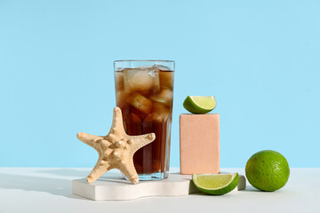 Glass of cold Cuba Libre cocktail with starfish and plaster podiums on white table against blue...