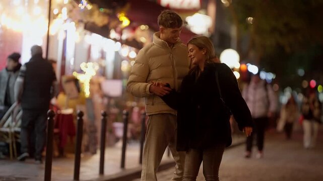 A young couple is holding hands on a Paris street at night The background features illuminated storefronts and other pedestrians The scene captures a moment of love and affection in an urban setting i