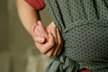 A close-up of a newborn tiny feet held in a parent hands while in a baby sling, emphasizing unity,...