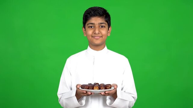 Portrait of a smiling young boy in a white thobe holding a plate of dates on a green background during ramadan.