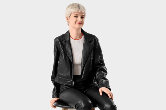Stylish young woman in leather jacket sitting on light background