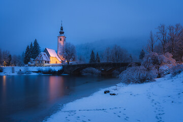Fototapeta premium Snowy church by Lake Bohinj in Slovenia reflecting calm winter evening atmosphere