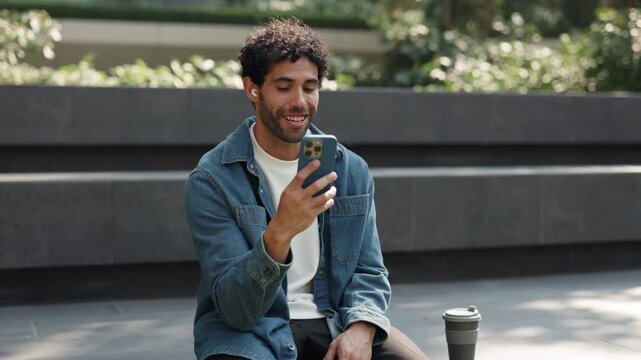 Portrait of positive Hispanic man talks on video call by smartphone as sits on parapet. Bearded Latin male in denim shirt greets by V-gesture on online chat via cellphone outdoors