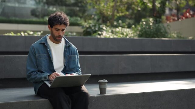 Portrait of young Hispanic man freelancer types on laptop as sits on park parapet. Bearded Latin male programmer develops software on portable computer resting on city street