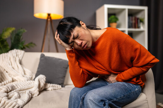 Asian woman suffering from stomach ache and headache on sofa. Healthcare and medicine concept. Japanese person with abdominal pain, nausea or menstrual cramps feeling unwell at home.