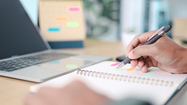 Business professional writing task notes in notebook beside laptop with colorful sticky notes. Concept of productivity, work planning, time management, organization and modern office workflow.