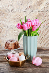 Golden bowl with pink Easter eggs on wooden table. Bouquet of tulips and traditional cake in the background.