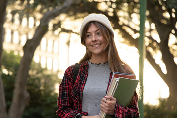 Female student smiling, holding green notebook stack, wearing beanie and backpack on campus