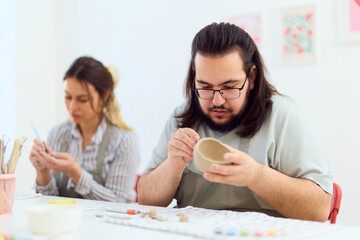 Fototapeta premium Friendly Pottery Class: Man Crafting Bowl While Woman Shapes Clay in Bright Studio