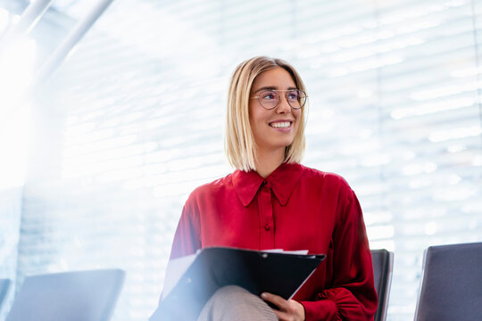 Smiling young businesswoman with folder sitting in waiting area